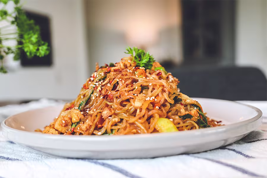 A plate of stir-fried noodles mixed with vegetables, shrimp, and garnished with parsley and sesame seeds sits on a striped cloth.