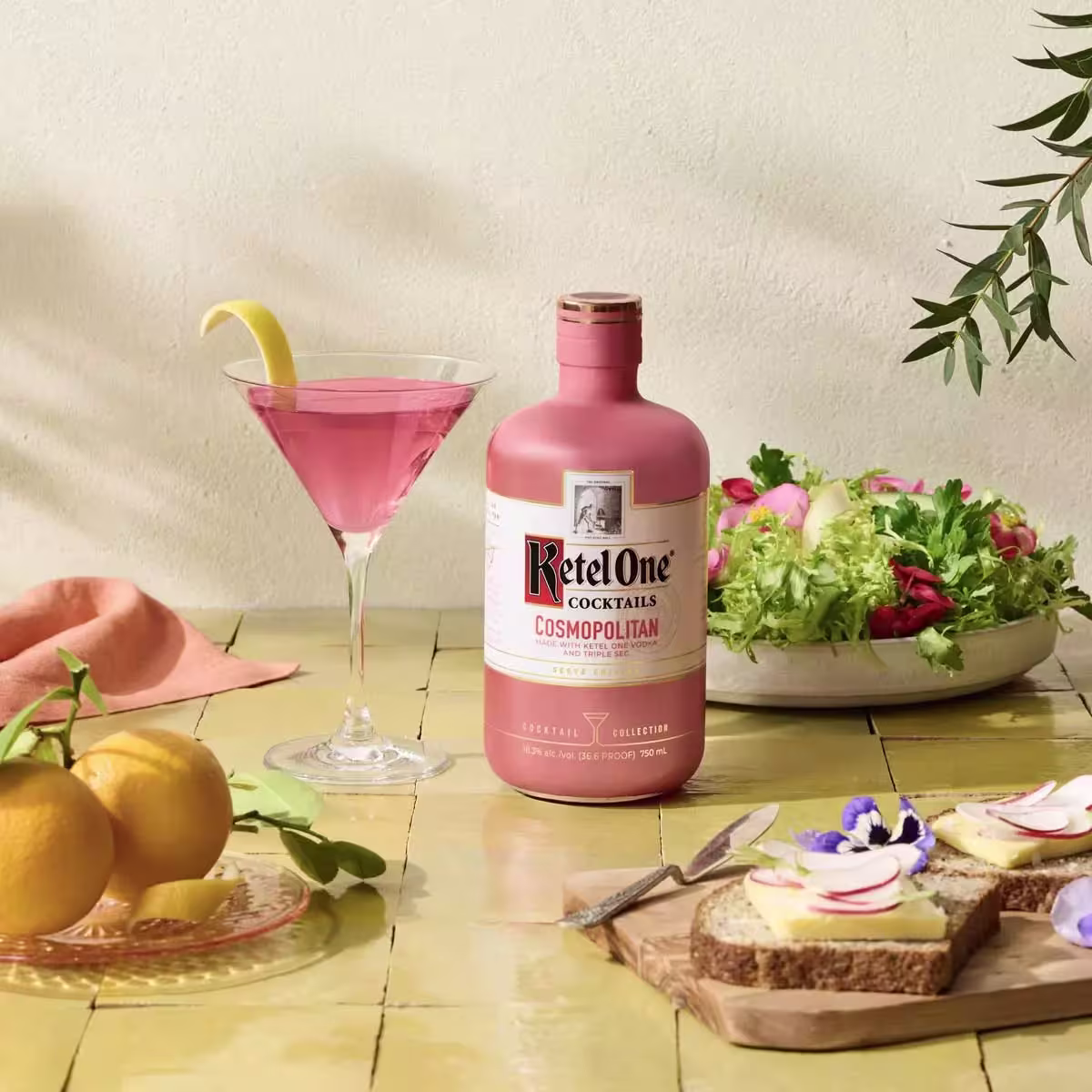 A bottle of Ketel One Cosmopolitan, a cocktail glass with a pink drink and lemon twist, bread slices with toppings, a salad bowl, and lemons on a yellow table.
