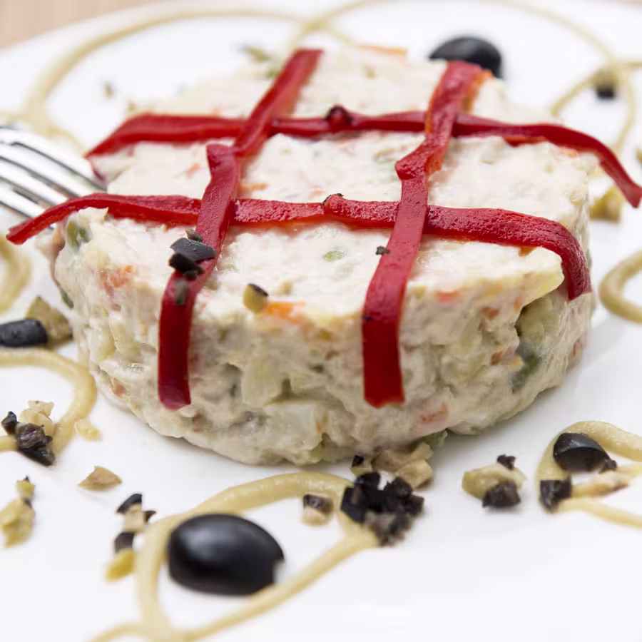 Ensaladilla rusa decorada con tiras de pimiento, aceitunas negras y alioli en Bienmesabe Príncipe de Vergara, un plato lleno de tradición y sabor.
