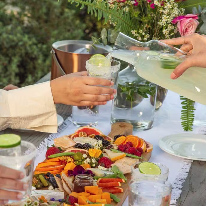 Hand pouring a light drink into a glass beside a colorful charcuterie board with fruits and flowers on an outdoor table.