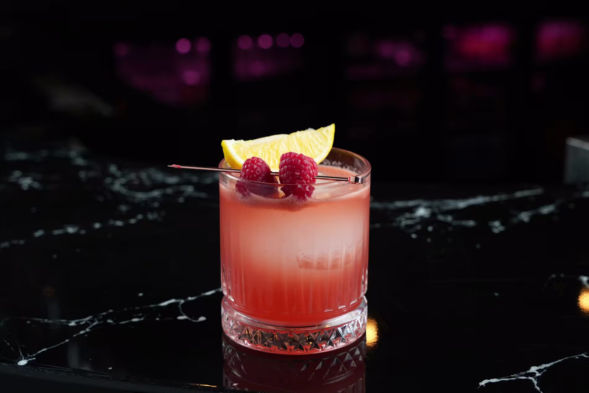 A pink cocktail in a glass garnished with two raspberries and an orange slice on a skewer, set on a black marble surface with a blurred dark background.