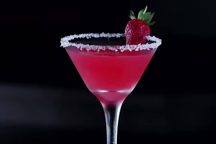 A red cocktail in a martini glass with a sugared rim, garnished with a whole strawberry, set against a dark background.