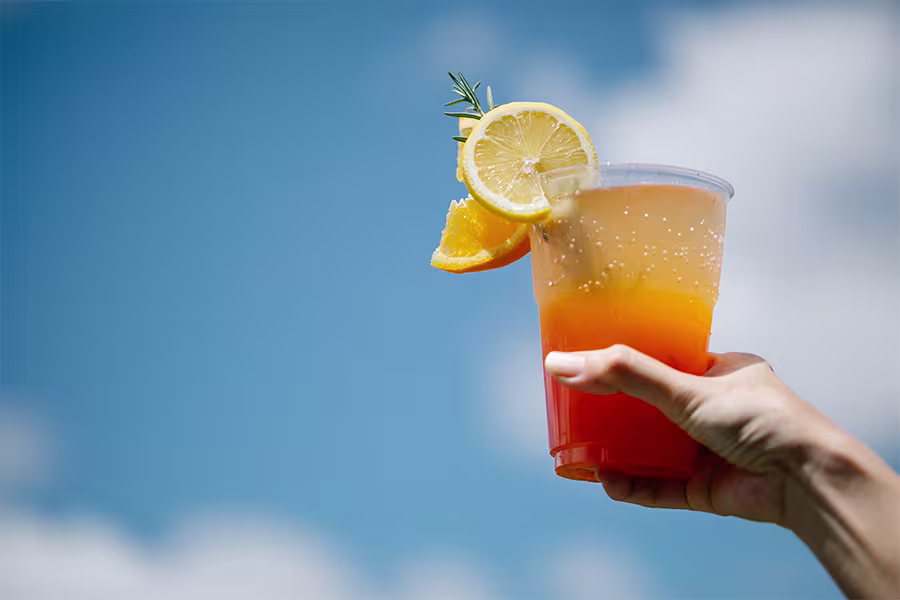 A hand holds up a plastic cup of orange and yellow layered drink garnished with a lemon slice, orange slice, and rosemary, against a blue sky with clouds.