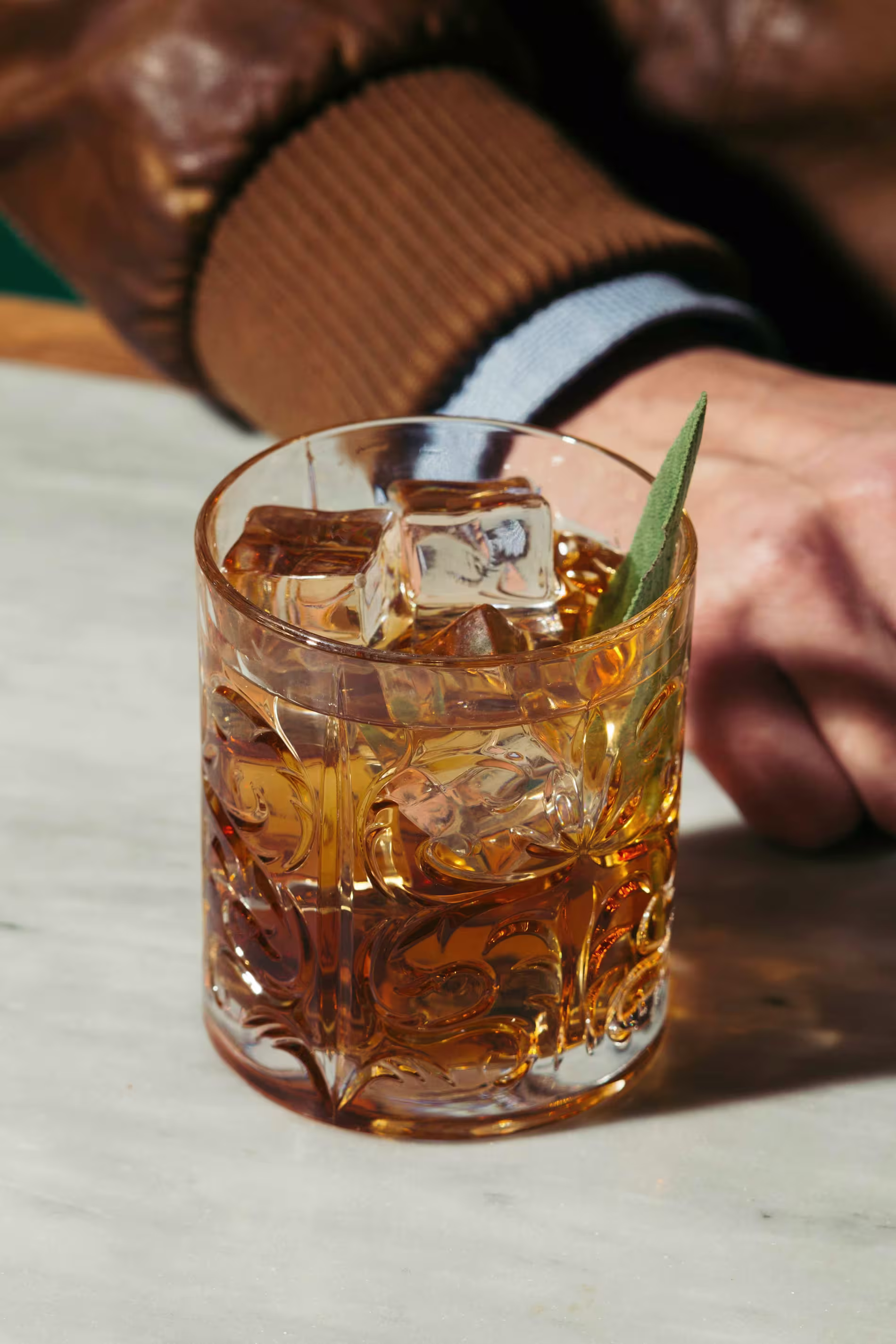 A glass of whiskey with ice cubes and a herb garnish sits on a marble surface next to a person wearing a brown jacket.