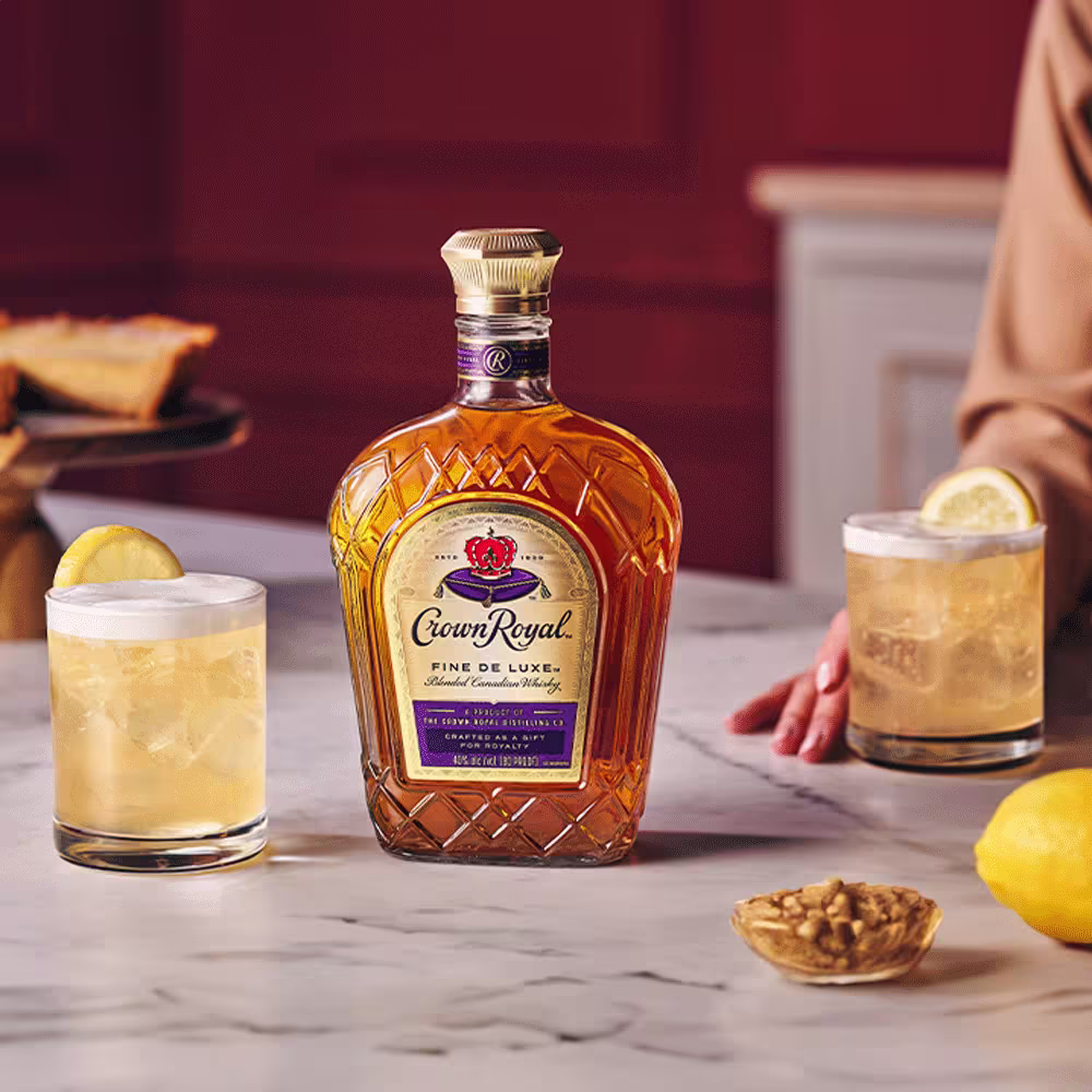 A bottle of Crown Royal whisky is displayed on a marble table between two cocktails garnished with lemon, with pie and walnuts in the background.