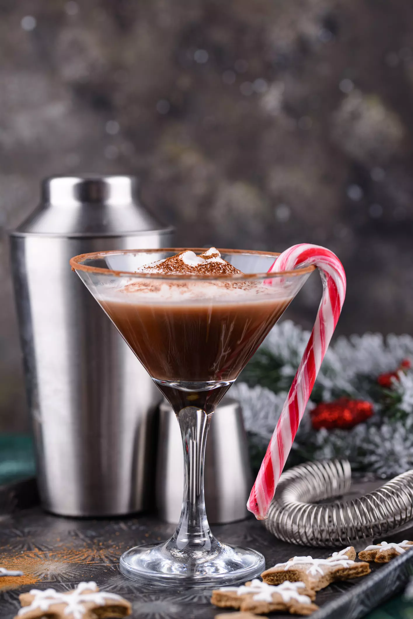 A martini glass filled with a chocolate cocktail, topped with whipped cream and cocoa powder, garnished with a candy cane. A cocktail shaker and festive decorations are in the background.