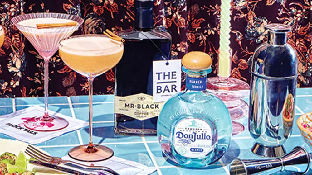 A table with cocktails in coupe glasses, a bottle of Don Julio tequila, Mr. Black coffee liqueur, a cocktail shaker, and various bar accessories.