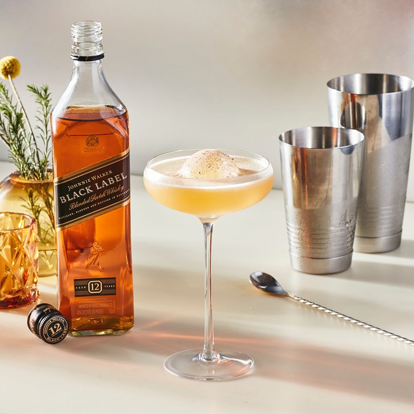 La imagen muestra una botella de whisky Johnnie Walker Black Label, una copa rellena con el cóctel Whisky Sour y otros elementos de coctelería.