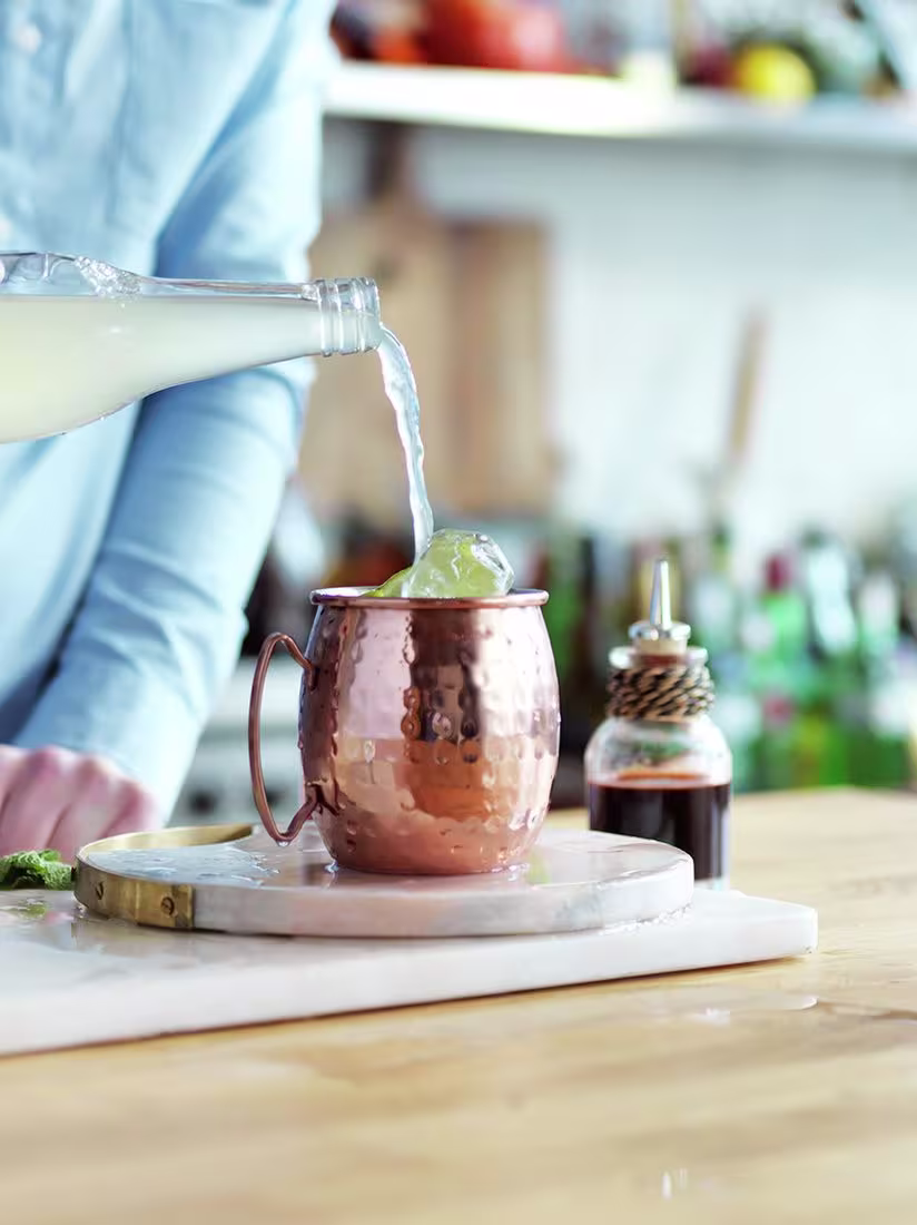 La imagen muestra el cóctel Moscow Mule en una taza de cobre, con una persona al fondo sirviendo zumo de limón en el vaso.