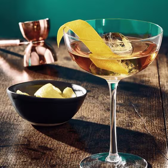 A glass of amber-colored cocktail with a large ice cube and a twist of lemon peel on a wooden surface.