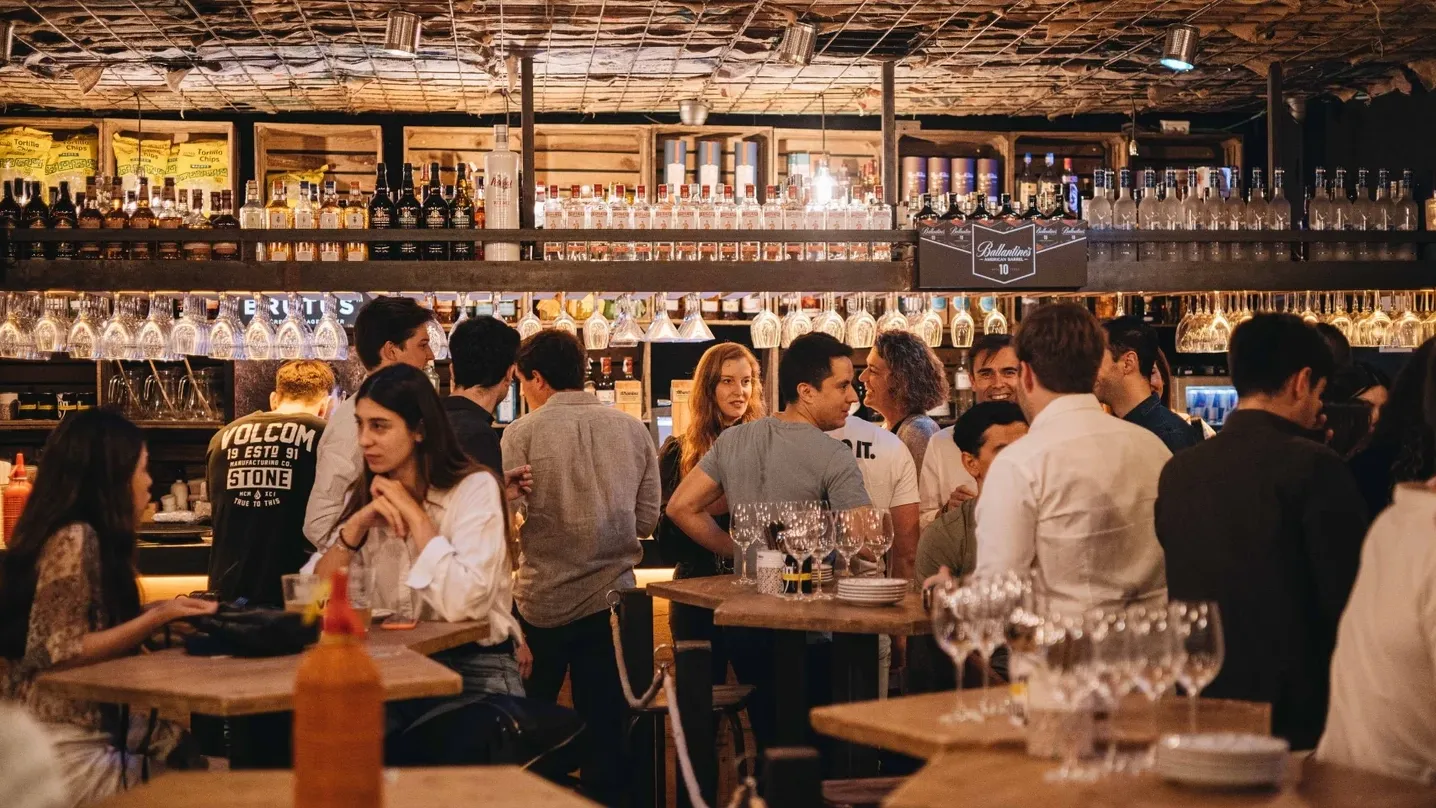 4 Latas Tuset: Bar repleto de gente, copas colgando sobre la barra, botellas en estanterías y mesas altas con platos y vasos. Ambiente animado con iluminación cálida y techo de malla.