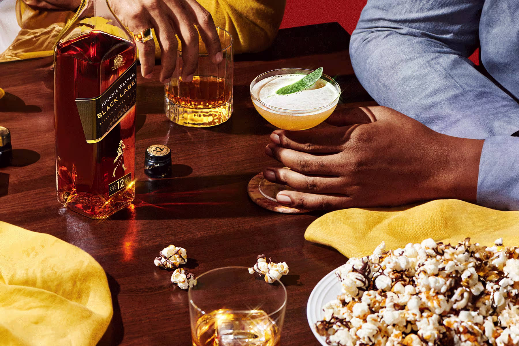 Two people sit at a table with a bottle of Johnnie Walker Black Label, two mixed drinks, a glass of whisky, and a plate of chocolate-drizzled popcorn.
