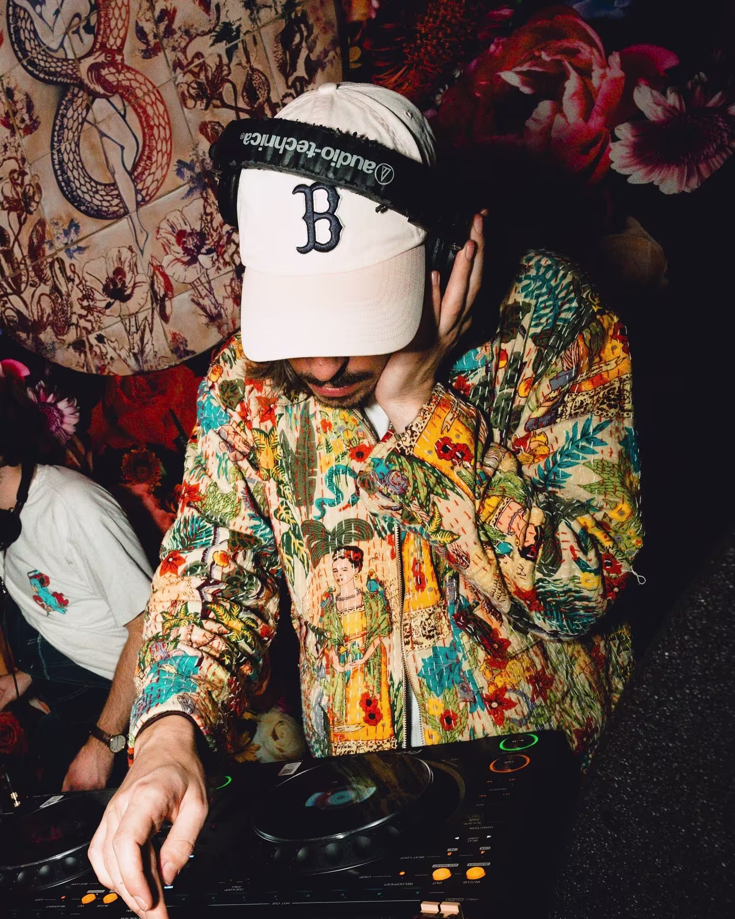 DJ en Los Amantes, con estilo vibrante, gorra blanca, camisa colorida y un fondo de estampados florales y artísticos, creando un ambiente único.