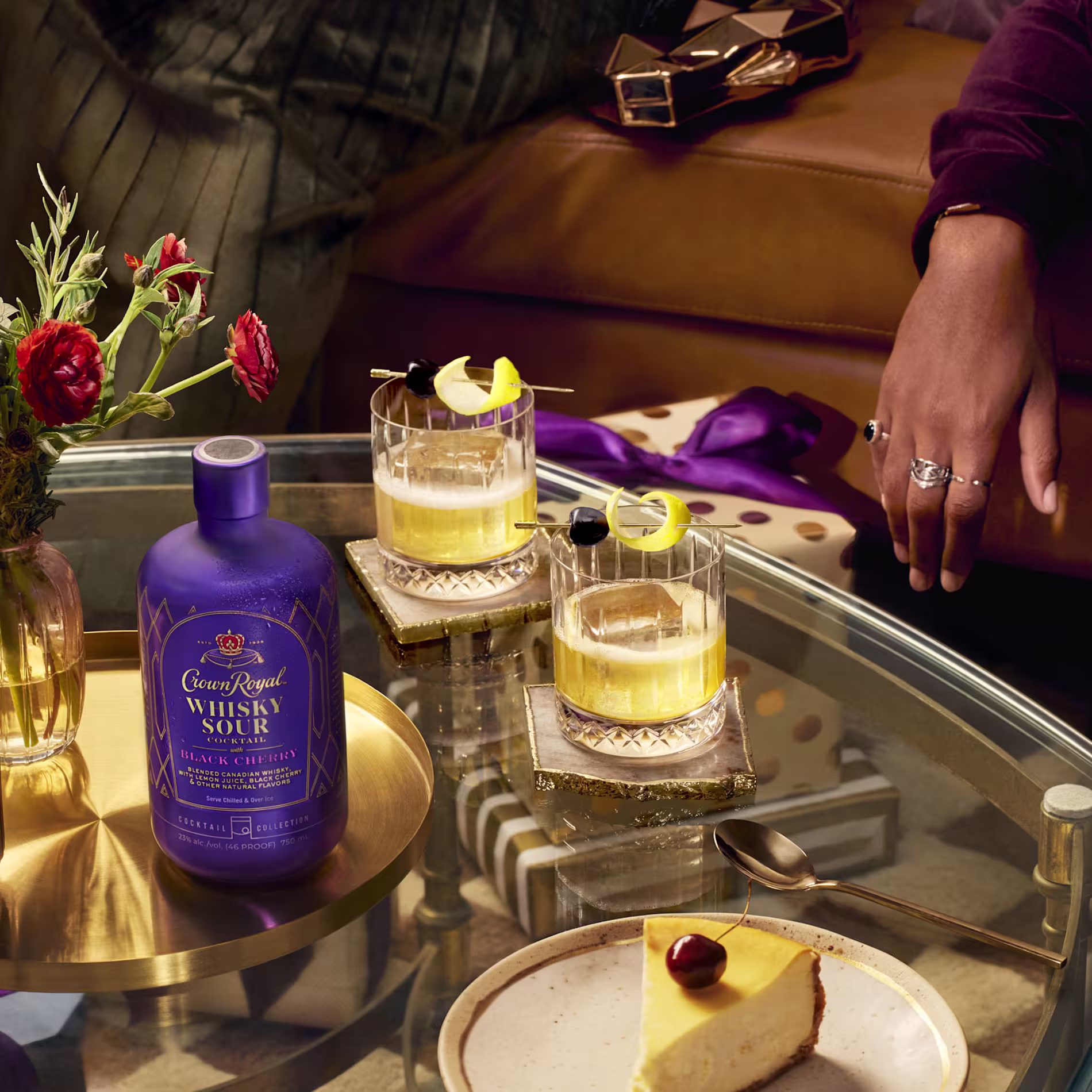 A bottle of Crown Royal Whisky Sour sits on a glass table with two whisky cocktails, a plate of cheesecake, flowers, and a hand resting on the couch nearby.
