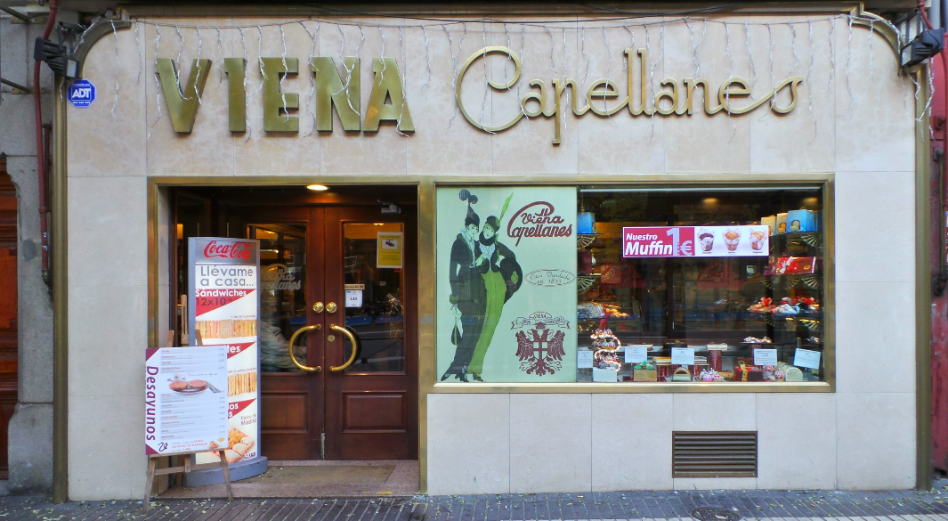 Fachada de Viena Capellanes en la calle Alcalá, con escaparate de bollería y carteles promocionales que invitan a desayunar o llevar productos a casa.