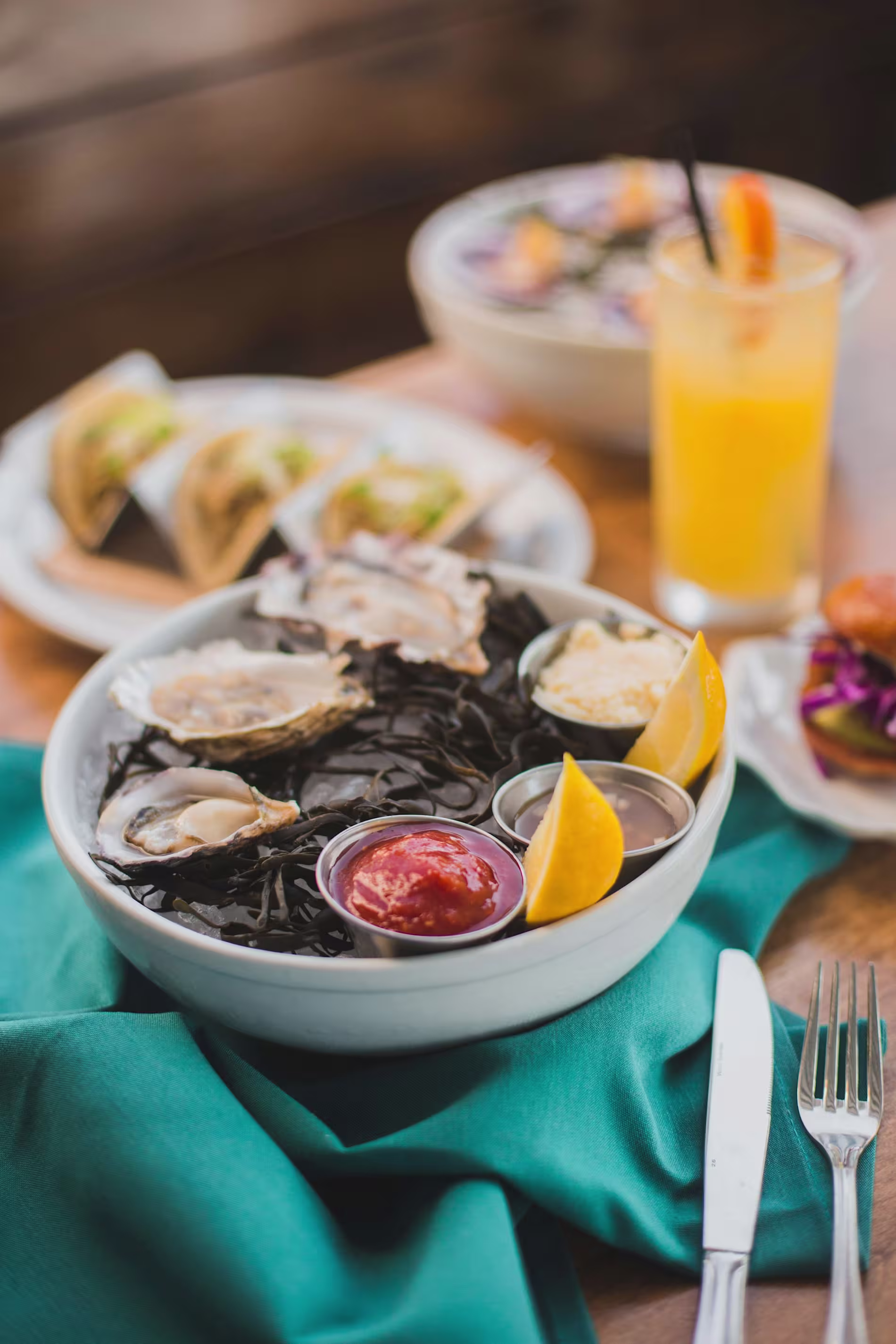 A bowl of oysters on a bed of seaweed with lemon slices and dipping sauces, placed on a table with a teal napkin, tacos, salad, and an orange drink in the background.