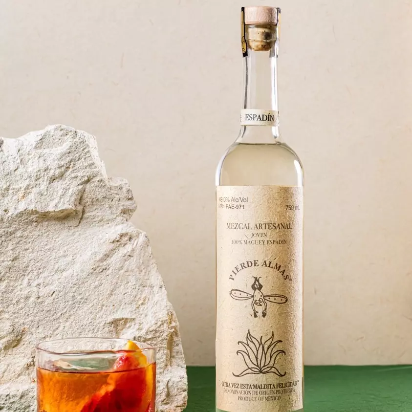A bottle of Pierde Almas mezcal stands upright beside a rock and a glass of cocktail garnished with an orange slice, set on a green surface against a light background.