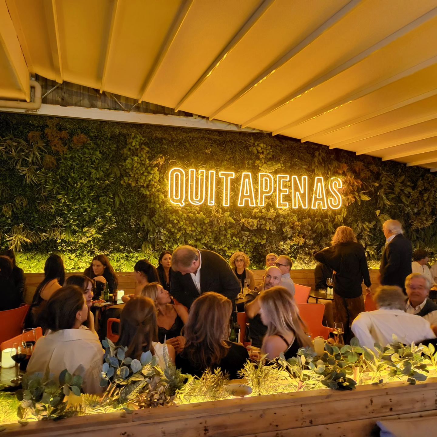 Quitapenas es un bar vibrante y acogedor, con una pared de plantas y un letrero de neón que destaca su nombre. Bajo una iluminación cálida, los clientes disfrutan de una velada animada y social.