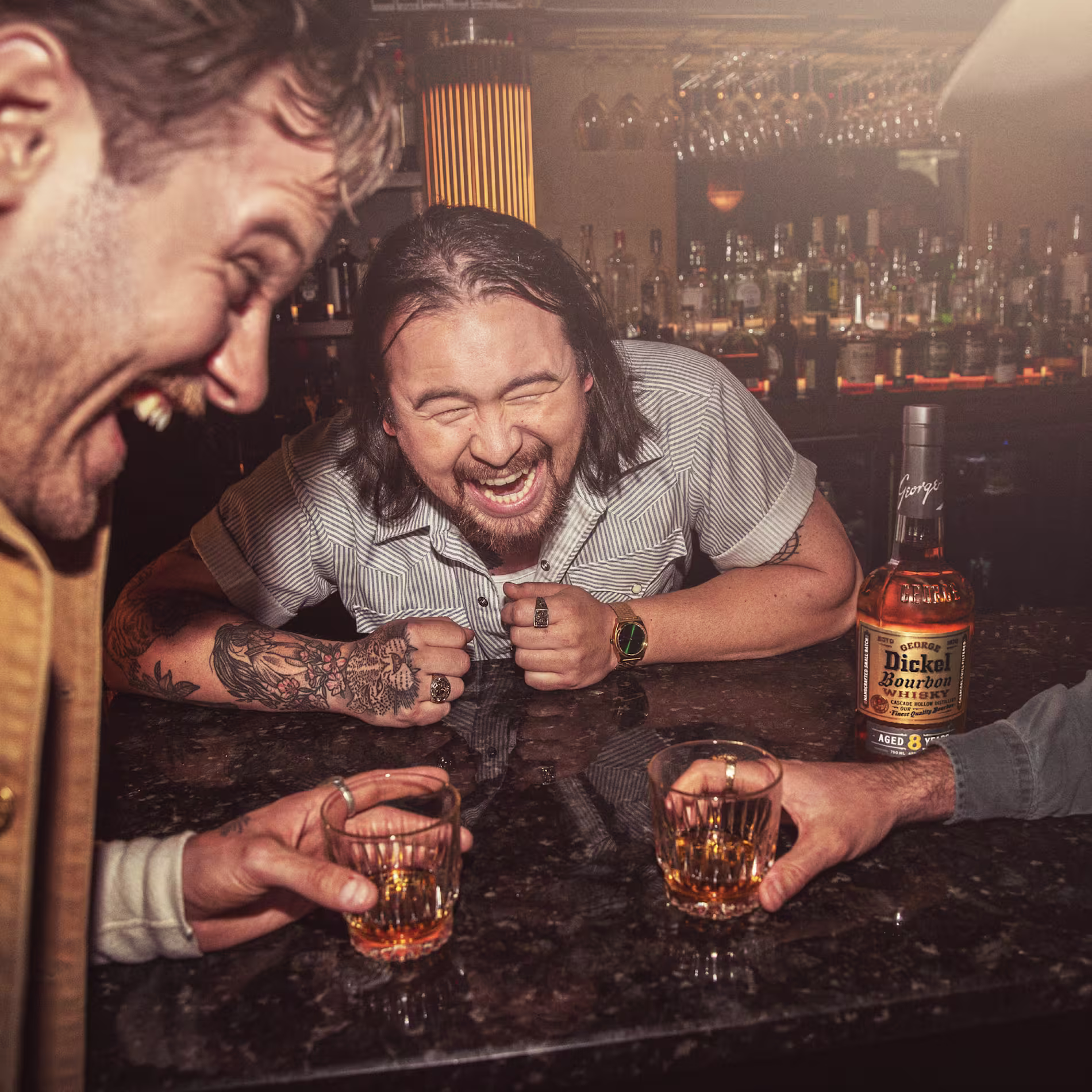 Three men laugh together at a bar, holding glasses of whiskey. A bottle of George Dickel Bourbon sits on the counter in front of them.