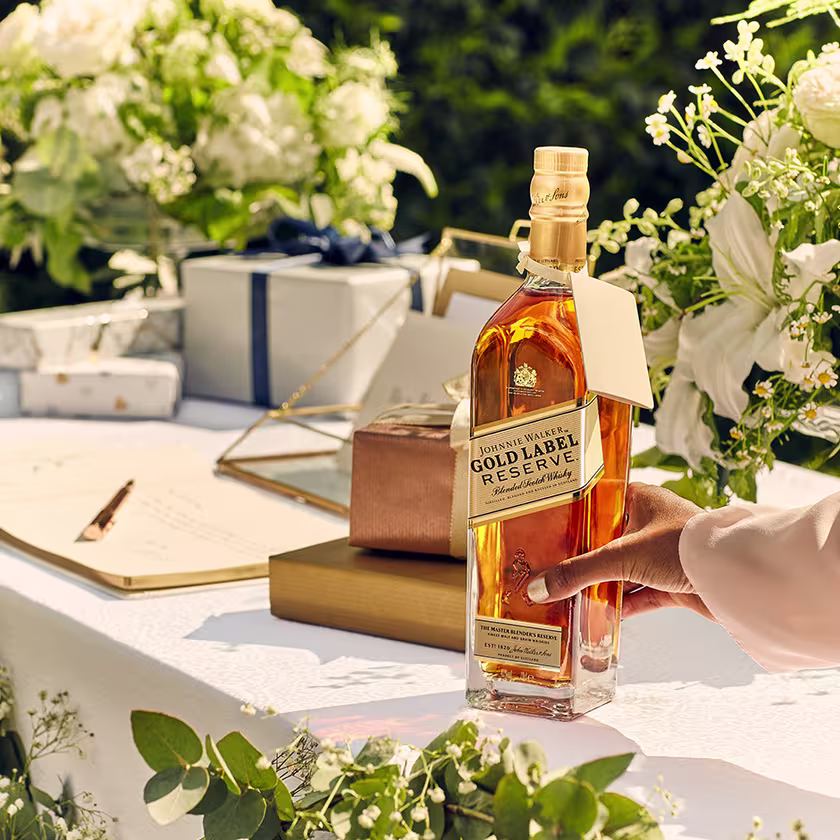 La imagen muestra la botella del whisky Johnnie Walker Gold Label Reserve con flores y regalos de fondo.