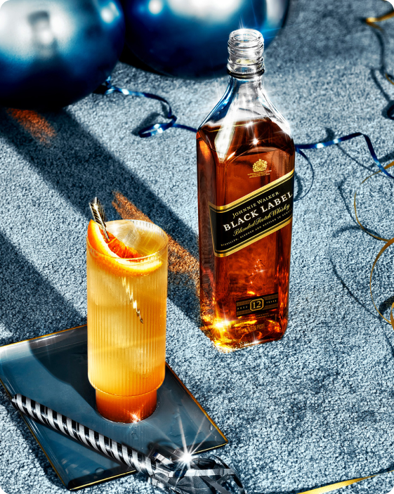 A bottle of Johnnie Walker Black Label whisky stands next to a cocktail garnished with an orange slice, set on a reflective tray on a blue textured surface.