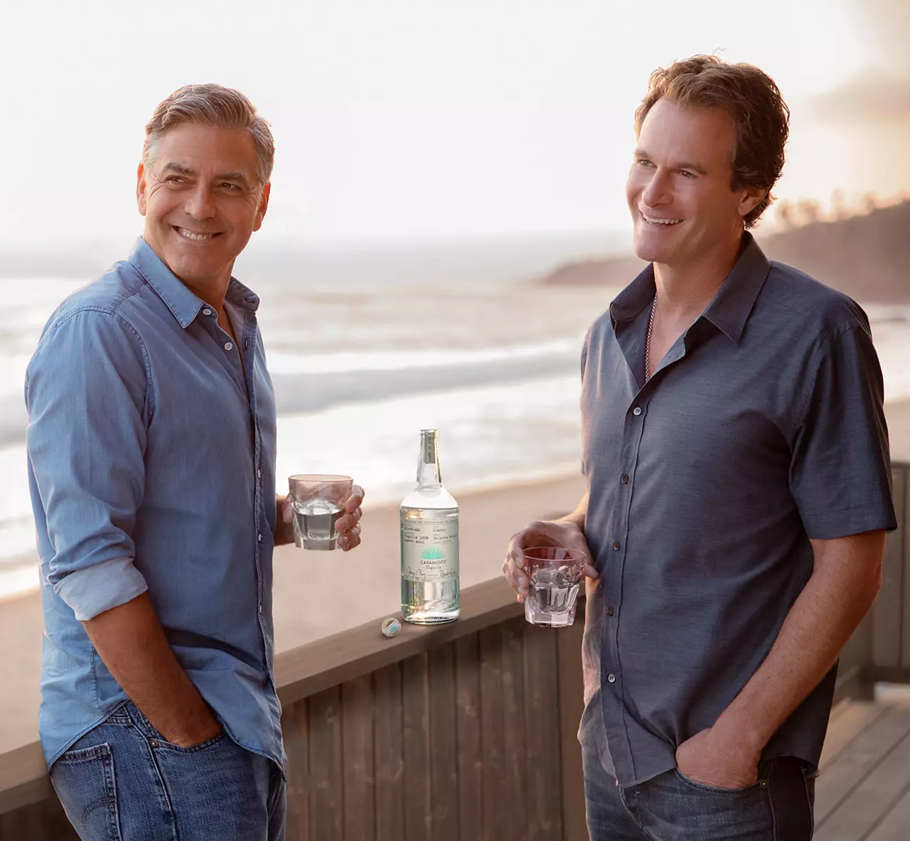 Two men stand outdoors by a wooden railing near the beach, each holding a glass with clear liquid. A large bottle of clear liquor is placed on the railing between them.