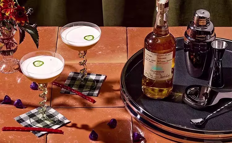 Two cocktails with foam and jalapeño slices, a bottle of Casamigos tequila, a cocktail shaker, measurement tools, and garnishes on a tiled table with a flower vase.