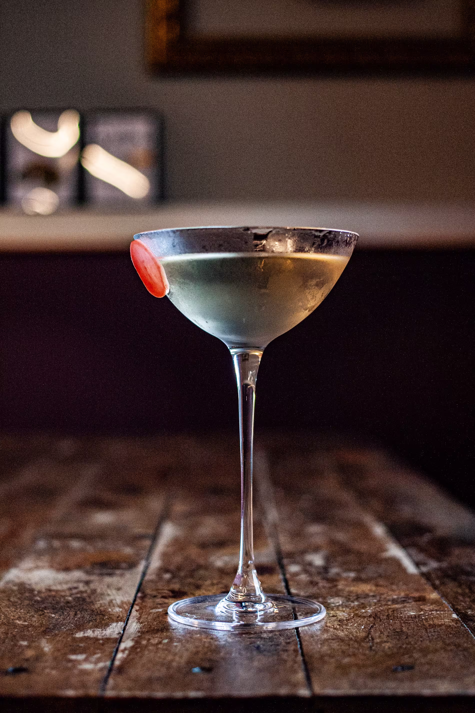 La imagen muestra una copa de martini con un líquido claro, posiblemente un cóctel, sobre una superficie de madera. Hay una rodaja de naranja en el borde de la copa. 