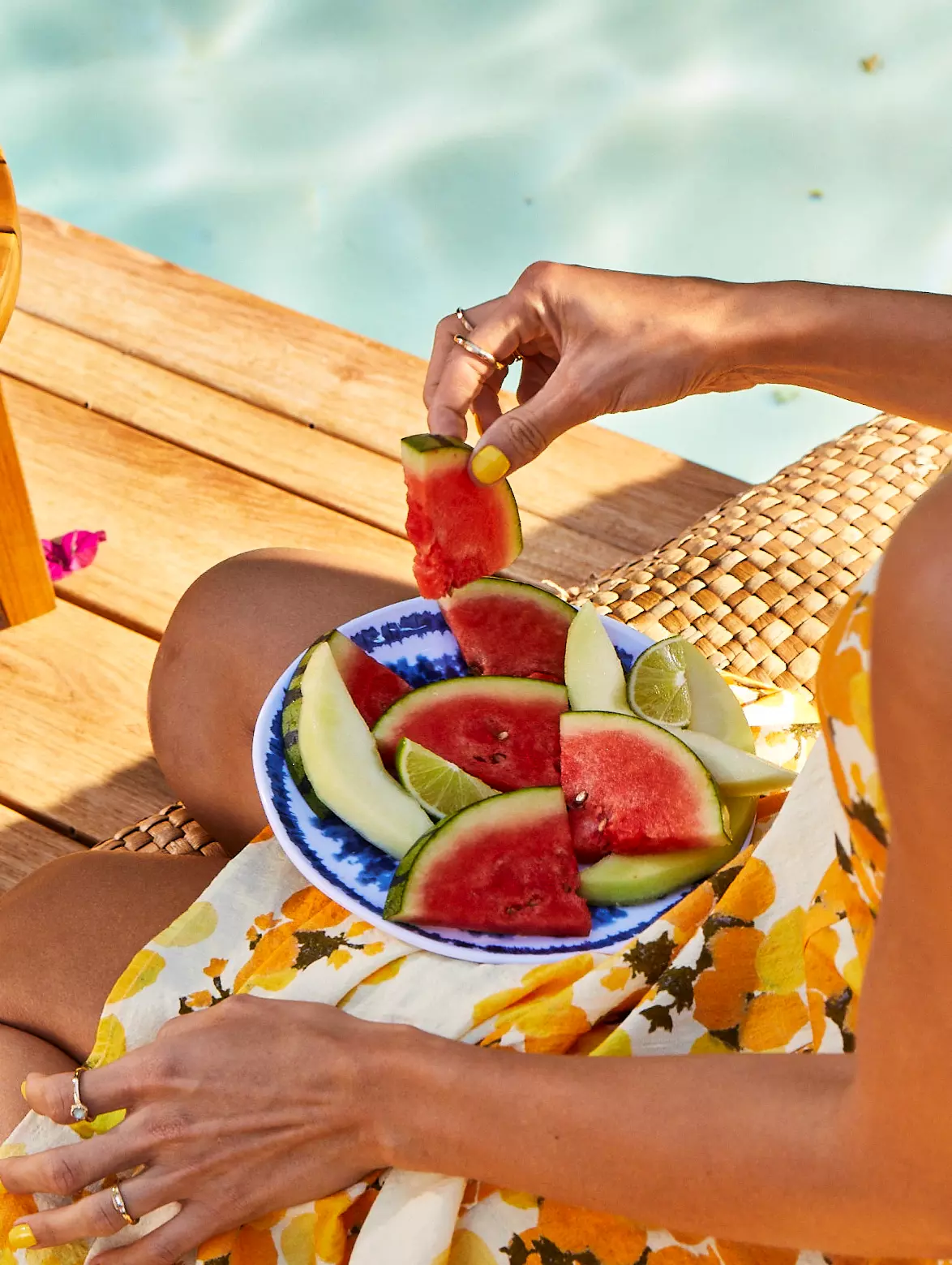 A person in a yellow floral dress holds a blue and white plate with watermelon and melon slices while sitting by a pool.