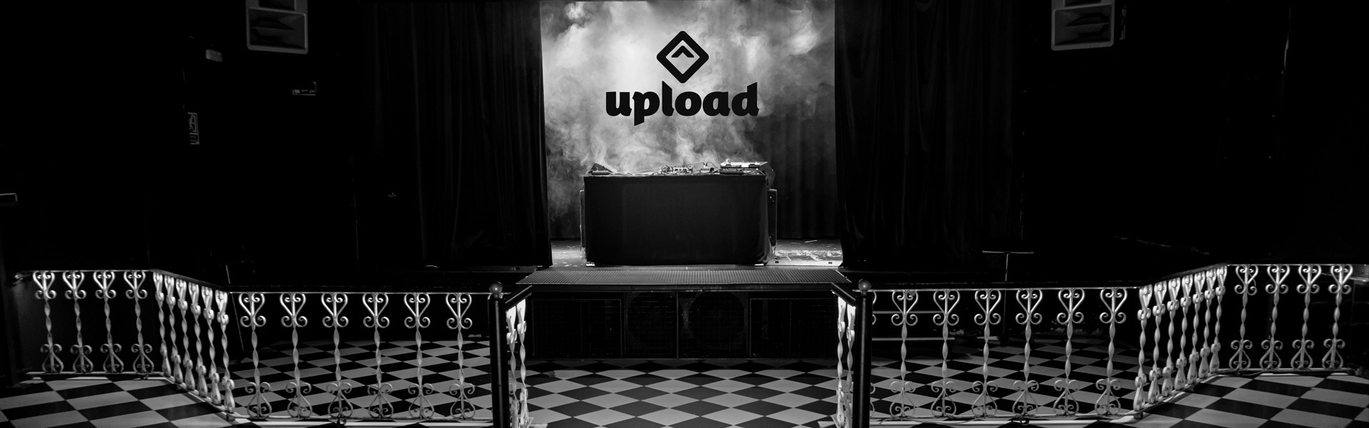 La foto muestra un entorno interior de la Sala Upload. Hay una gran pantalla en el escenario que muestra la palabra “upload” . El escenario está flanqueado por dos barricadas con un patrón de cuadros.