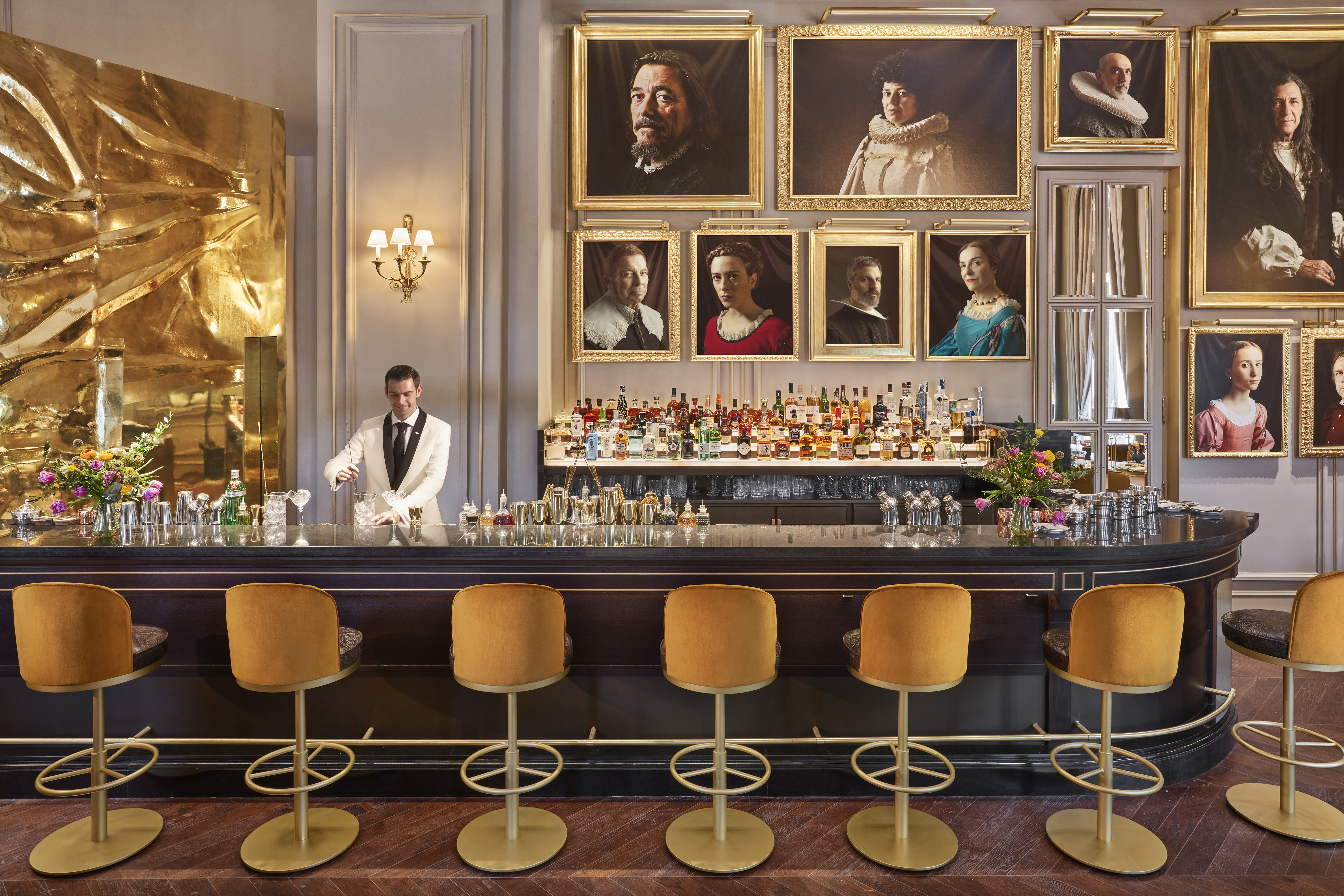 La foto muestra un elegante bar, el Pictura. Hay un camarero detrás del mostrador, que está bien surtido con varias botellas de alcohol. El mostrador es negro, curvado y tiene un acabado brillante. Frente al mostrador, hay varios taburetes altos.