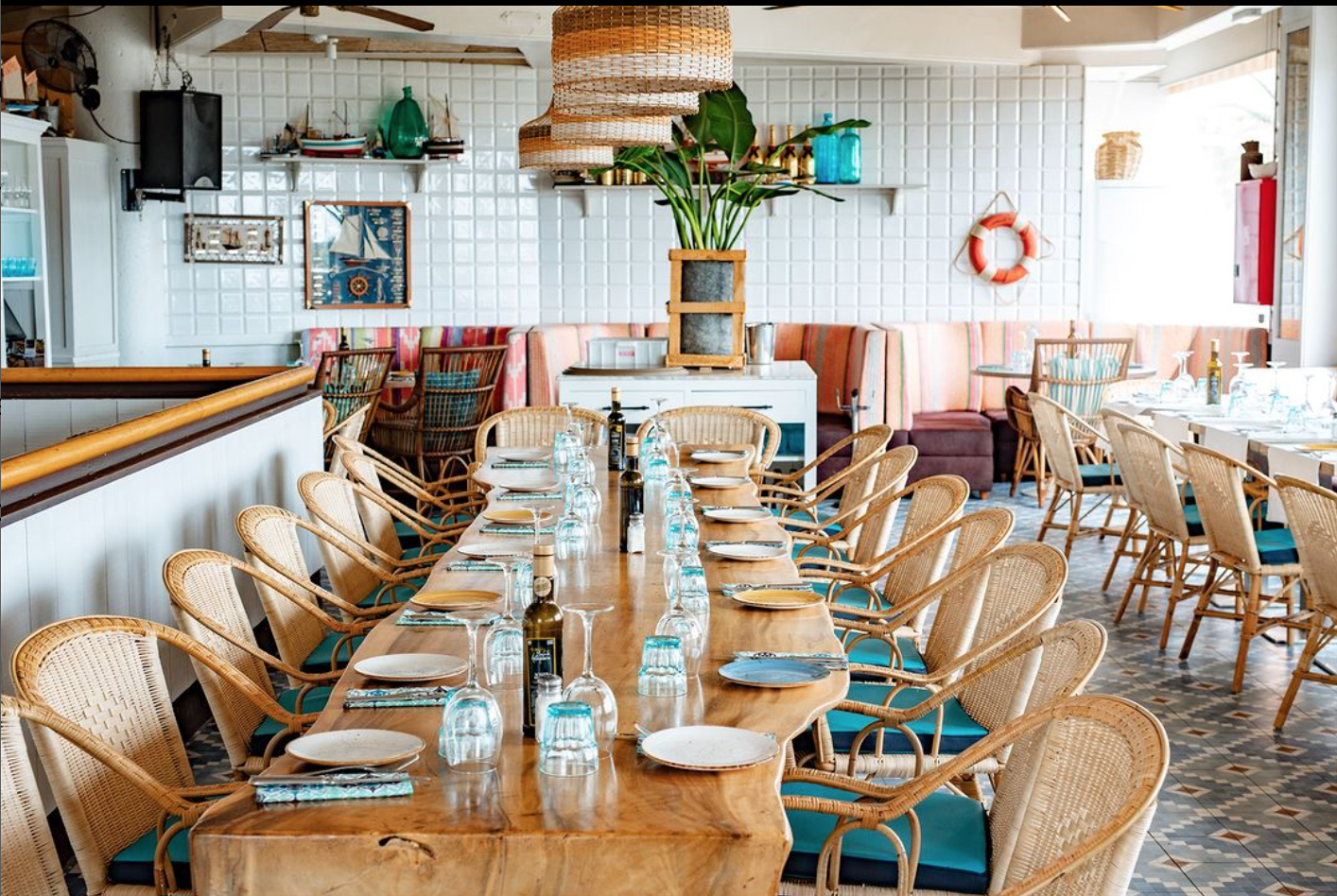 La imagen muestra el interior de un área de comedor de el Marina Bay. El entorno incluye una larga mesa de madera con sillas de ratán alrededor, dispuestas con platos, cubiertos y vasos. La decoración tiene un tema costero o náutico