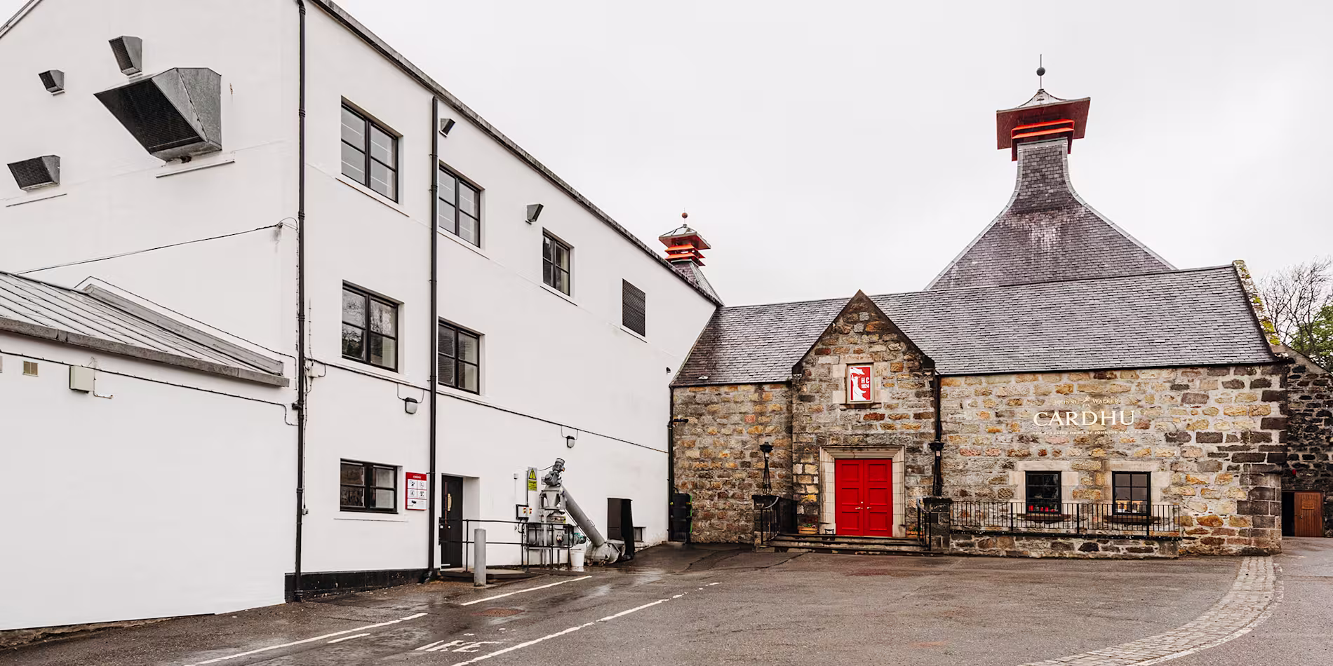 El edificio de piedra marrón de la destilería Cardhu está situado junto a un edificio blanco. El edificio tiene un tejado inclinado de pizarra y una puerta roja. Adosado al muro de piedra marrón hay un letrero metálico dorado con las palabras "Cardhu".