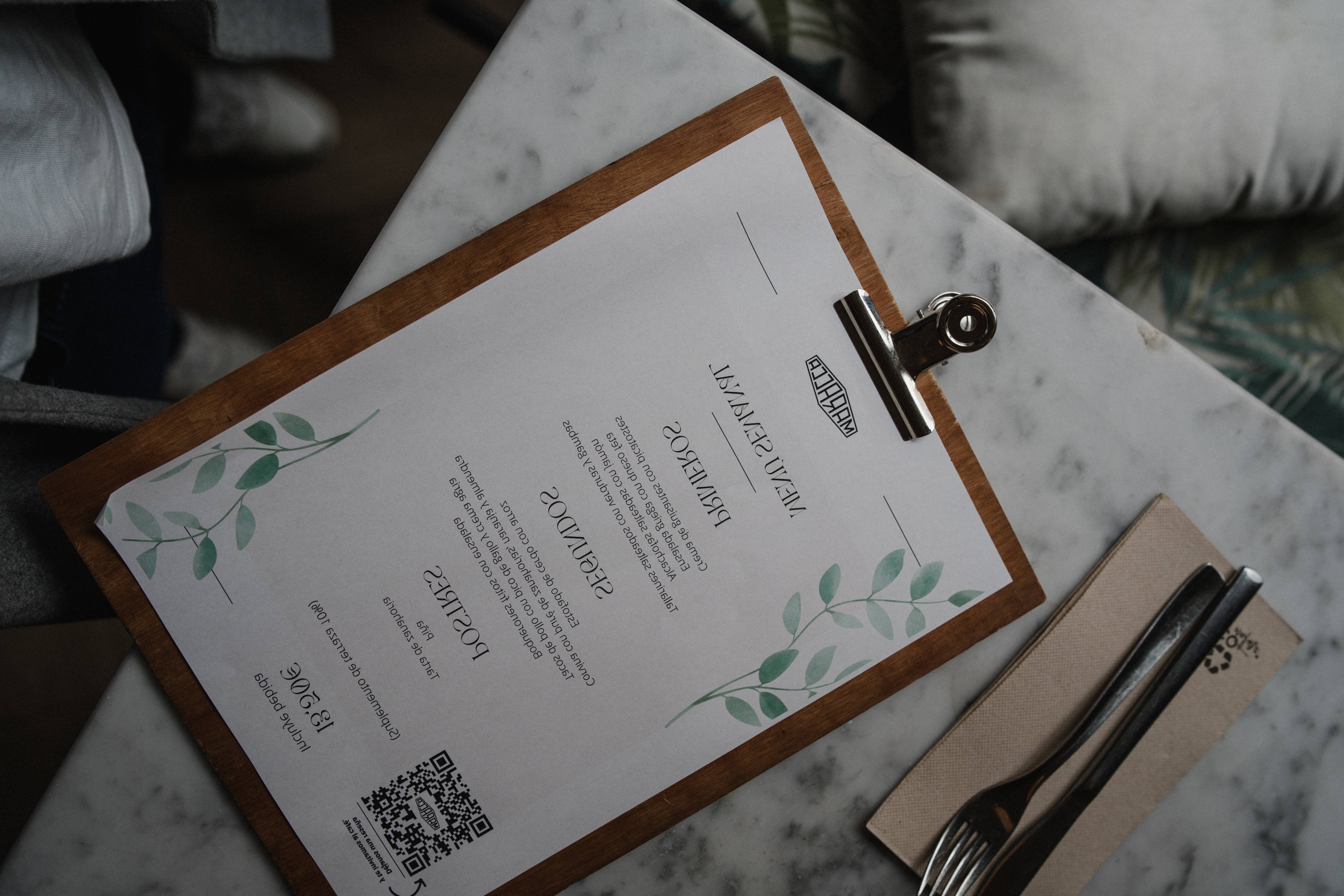 La imagen muestra un menú impreso colocado sobre una superficie de madera. La invitación incluye texto, algunos elementos decorativos como ramas con hojas, y un código QR en la esquina superior izquierda.
