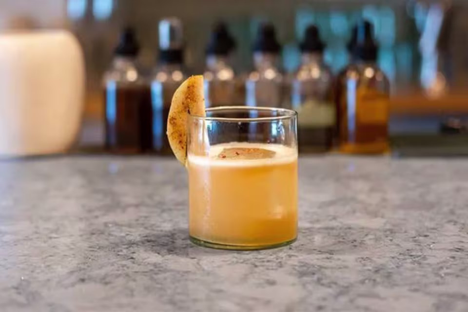 A short glass of orange cocktail with a foamy top and a spiced citrus garnish sits on a marble countertop; bottles are blurred in the background.