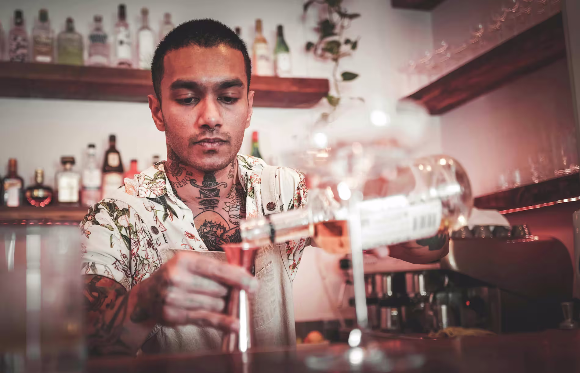 A bartender with tattoos pours a drink into a measuring jigger behind a bar stocked with bottles and glassware.
