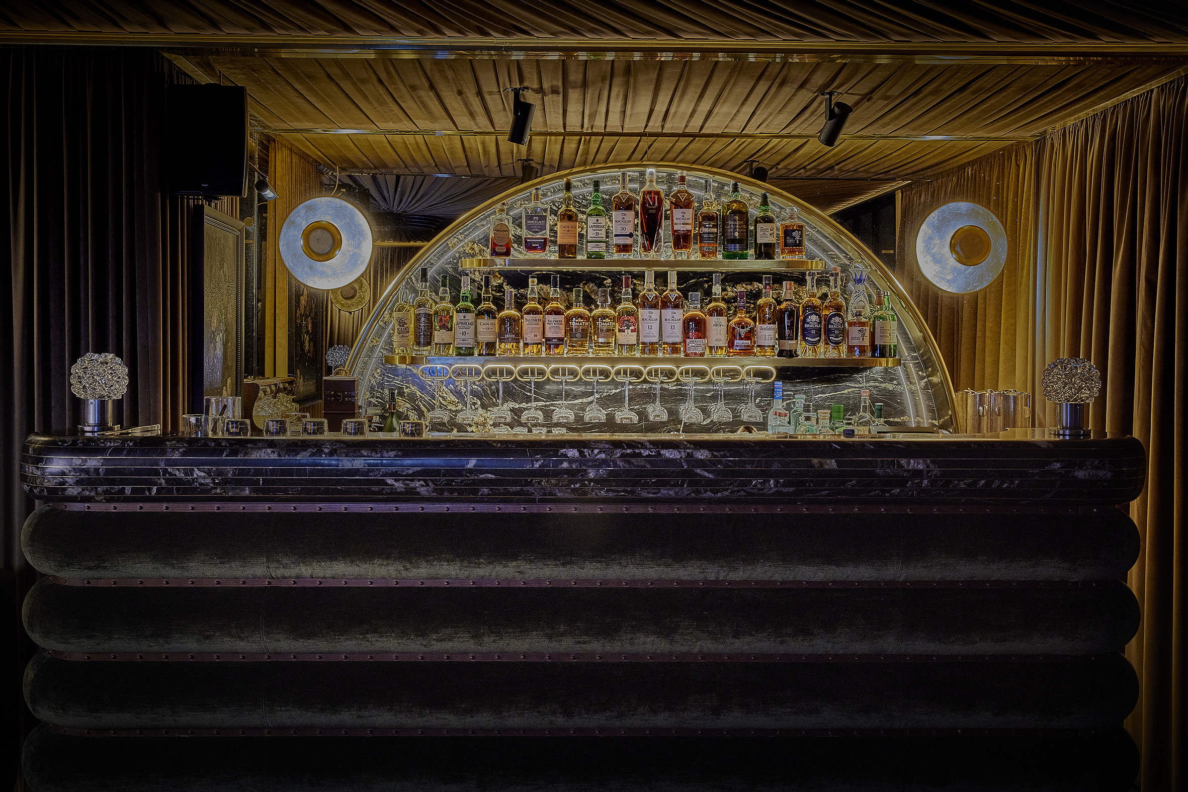 La imagen muestra el interior de El Coleccionista. La barra tiene una unidad de estanterías semicircular y escalonada para las botellas, con iluminación que resalta las botellas y la cristalería.
