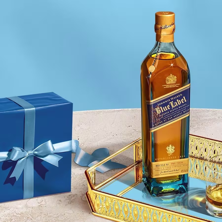 Una botella de whisky Johnnie Walker Blue Label sobre una bandeja dorada junto a un vaso y una caja de regalo azul con un lazo sobre un fondo azul claro.