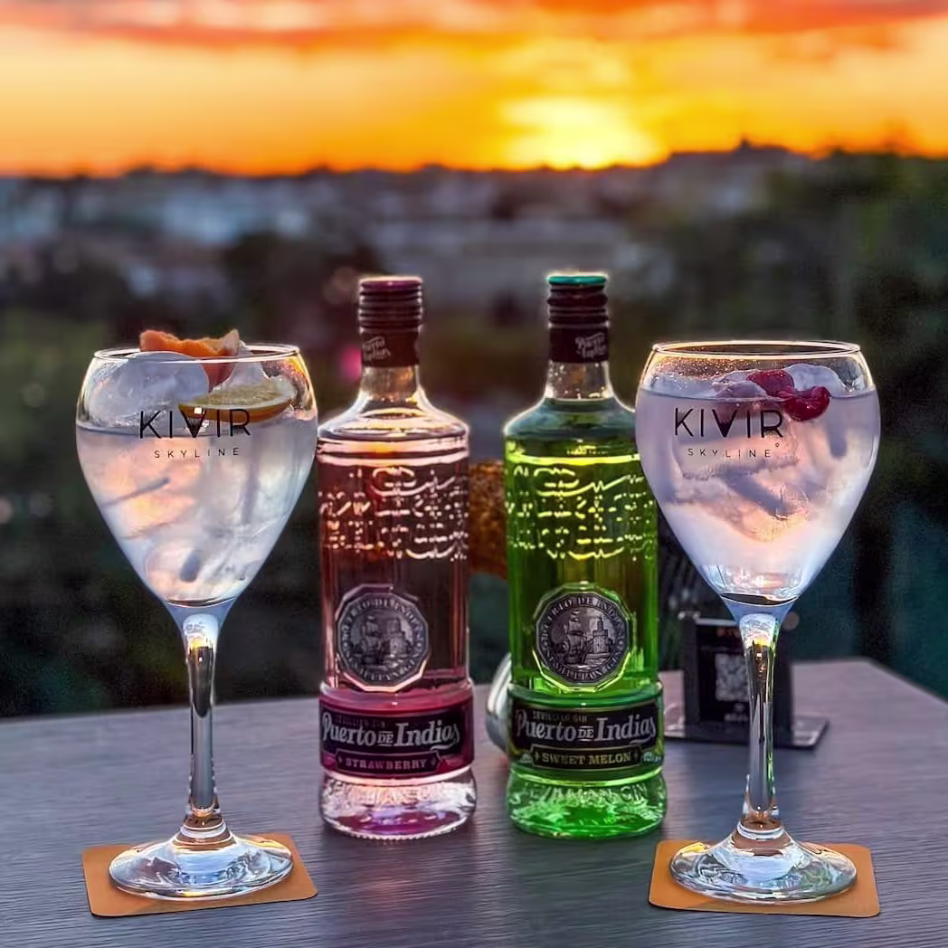 Kivir Skyline al atardecer, con dos copas de gin tonic frente a un cielo naranja y botellas de Puerto de Indias en primer plano.