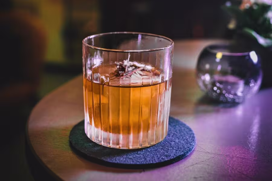 A glass of amber-colored drink with ice and garnish sits on a dark coaster atop a round table.