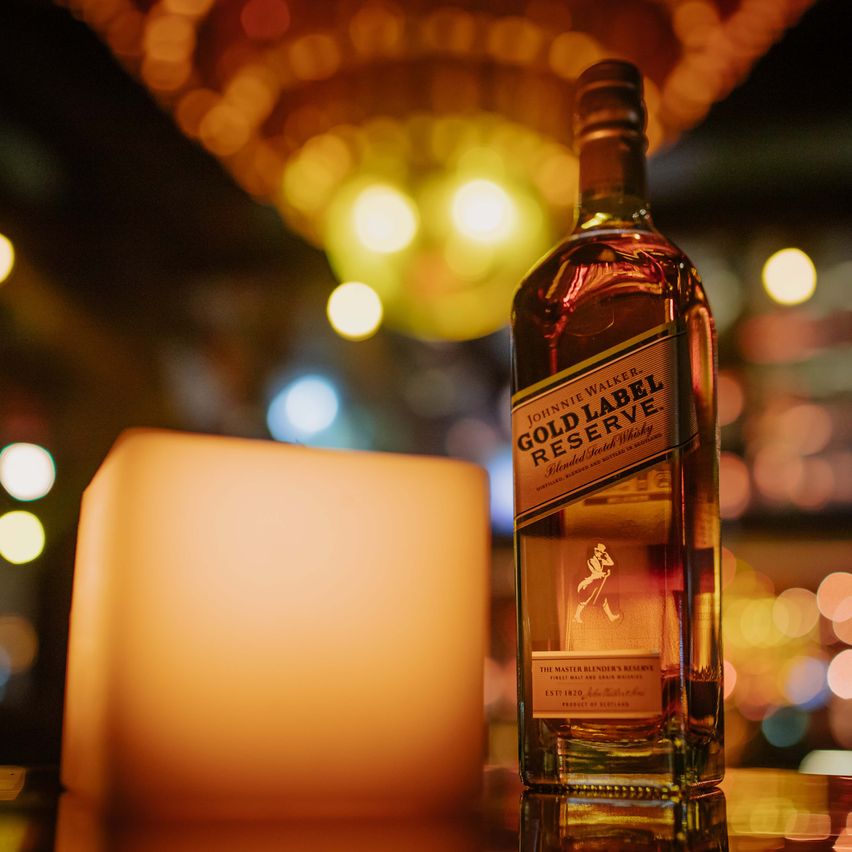 En la imagen se ve una botella de whisky Johnnie Walker Gold Label Reserve apoyado sobre una mesa y, a su lado, una vela. El ambiente es elegante y sofisticado.