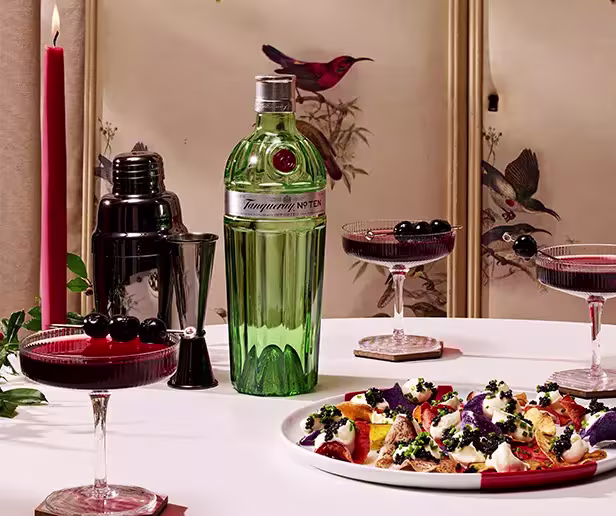 A bottle of Tanqueray gin, a cocktail shaker, three cocktails with cherries, a red candle, and a plate of food are arranged on a table with a bird-themed screen in the background.