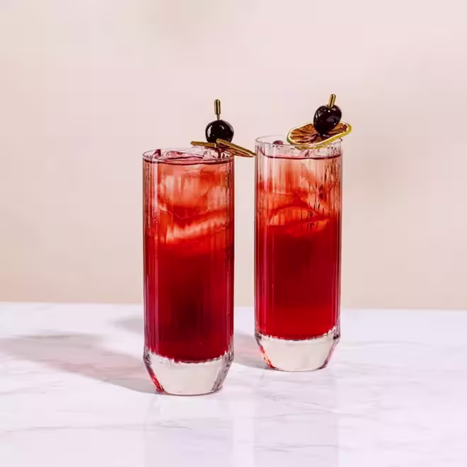 Two tall glasses filled with a red cocktail over ice, each garnished with a cherry and citrus slice on a skewer, set on a white marble surface.
