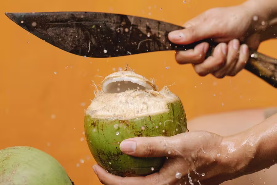 A person uses a large knife to cut open a green coconut, with coconut water splashing against an orange background.