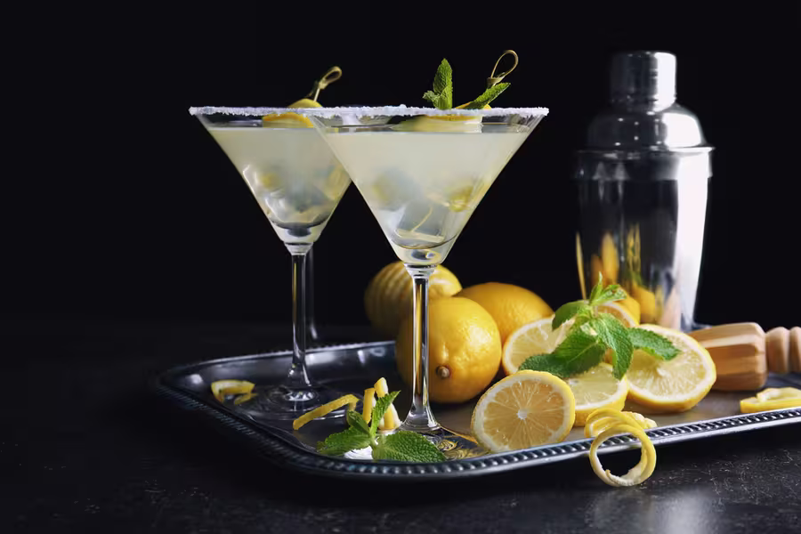 Two martini glasses with lemon cocktails and ice are on a tray, surrounded by fresh lemons, lemon slices, mint, a cocktail shaker, and a wooden juicer.