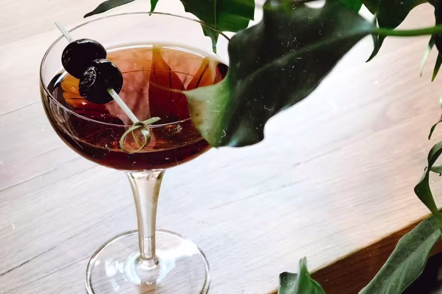 A cocktail in a coupe glass garnished with two dark cherries on a skewer, set on a light wooden surface near green leaves.