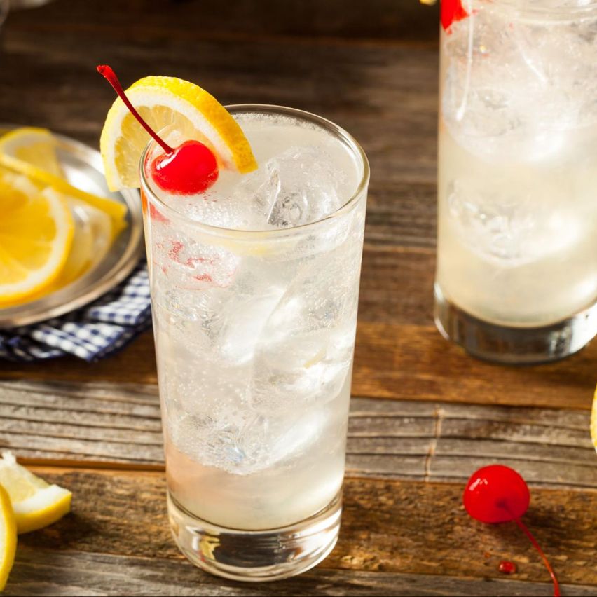 Imagen del trago Tom Collins, en dos vasos largos con hielo, decorados con una rodaja de limón y una cereza. Sobre una mesa de madera, con rodajas de limón alrededor