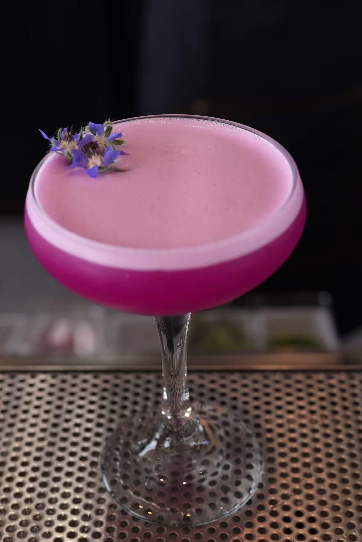A coupe glass filled with a vibrant pink cocktail topped with a layer of foam and garnished with small purple edible flowers, placed on a perforated metal surface.