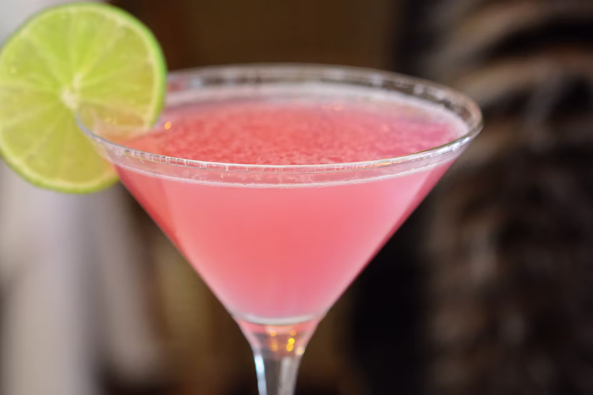 A pink cocktail served in a martini glass with a slice of lime on the rim.