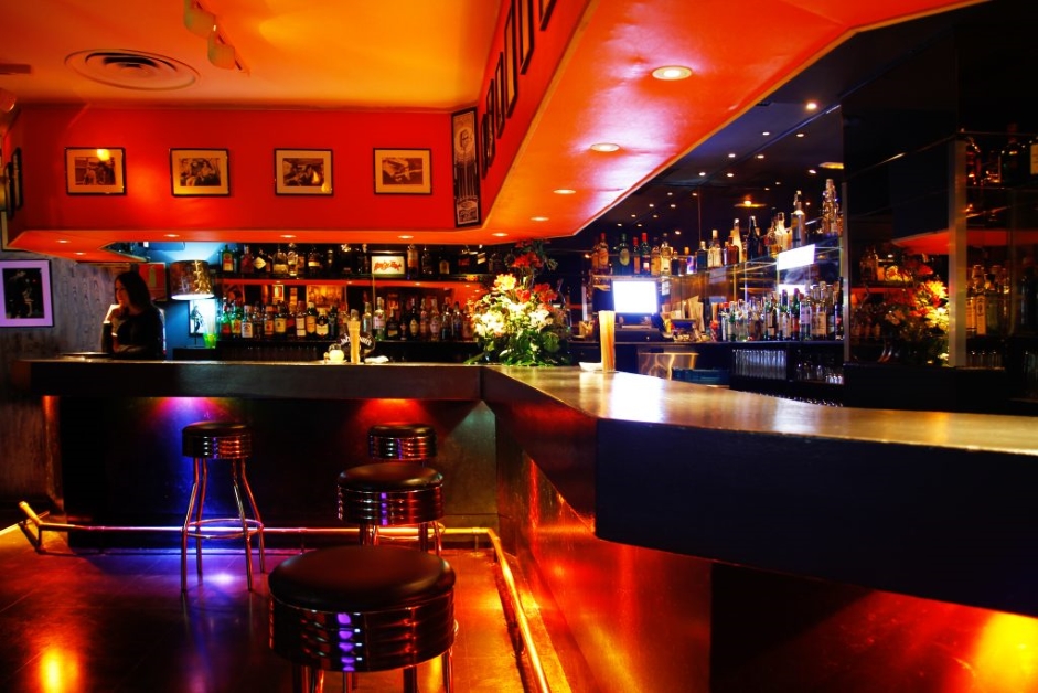 La foto muestra el interior del Honky Tonk. La zona del bar está iluminada con una luz cálida y ambiental que resalta la variedad de botellas en la estantería trasera. Hay varios taburetes negros con bases cromadas frente al mostrador del bar.